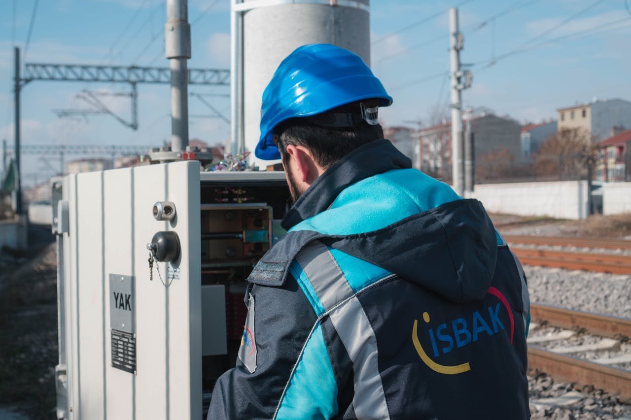 Instalaciones eléctricas en Valencia. An engineer in a blue hard hat examines a fuse box on a railway track outdoors.